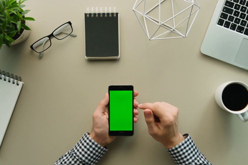 Man using smartphone with green screen on office desk background. Male  hands tapping on touchscreen, swiping, scrolling pages on cell phone. Tip  view.