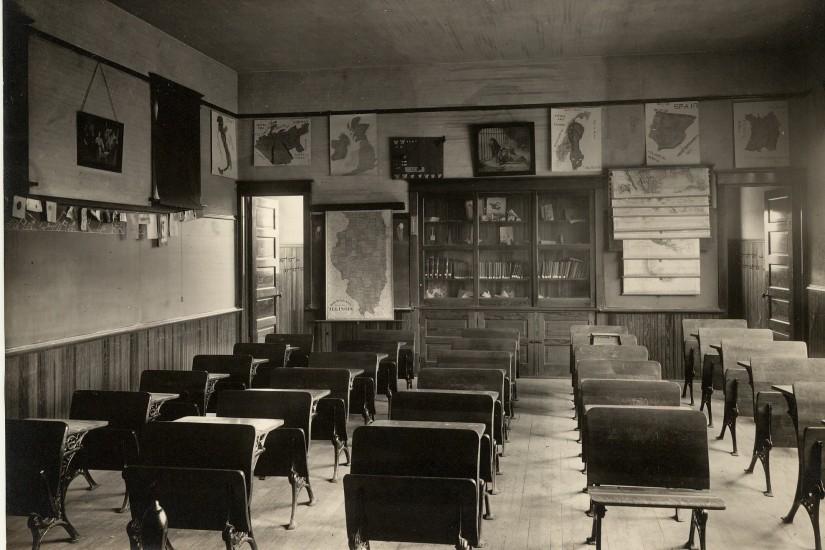This photograph, taken from the front of the classroom in Love School,  shows desks in rows, a book case, and maps on the walls.