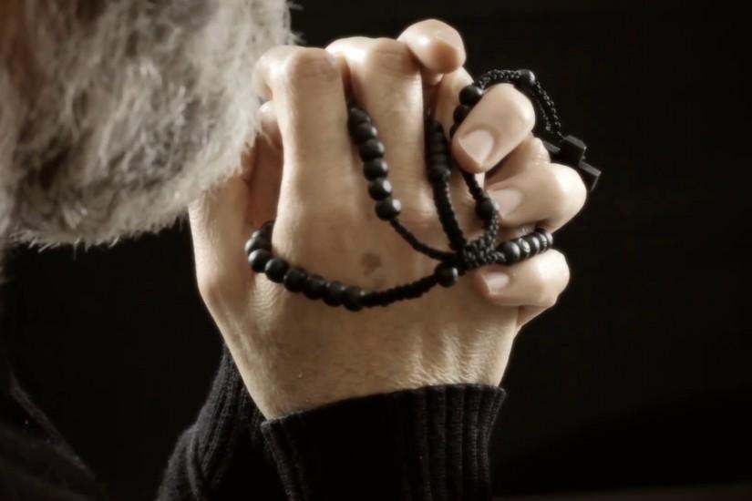 A faithful man, a believer, praying God with a rosary in his old wrinkled  hands, whispering a prayer. Profile shot. Black background.